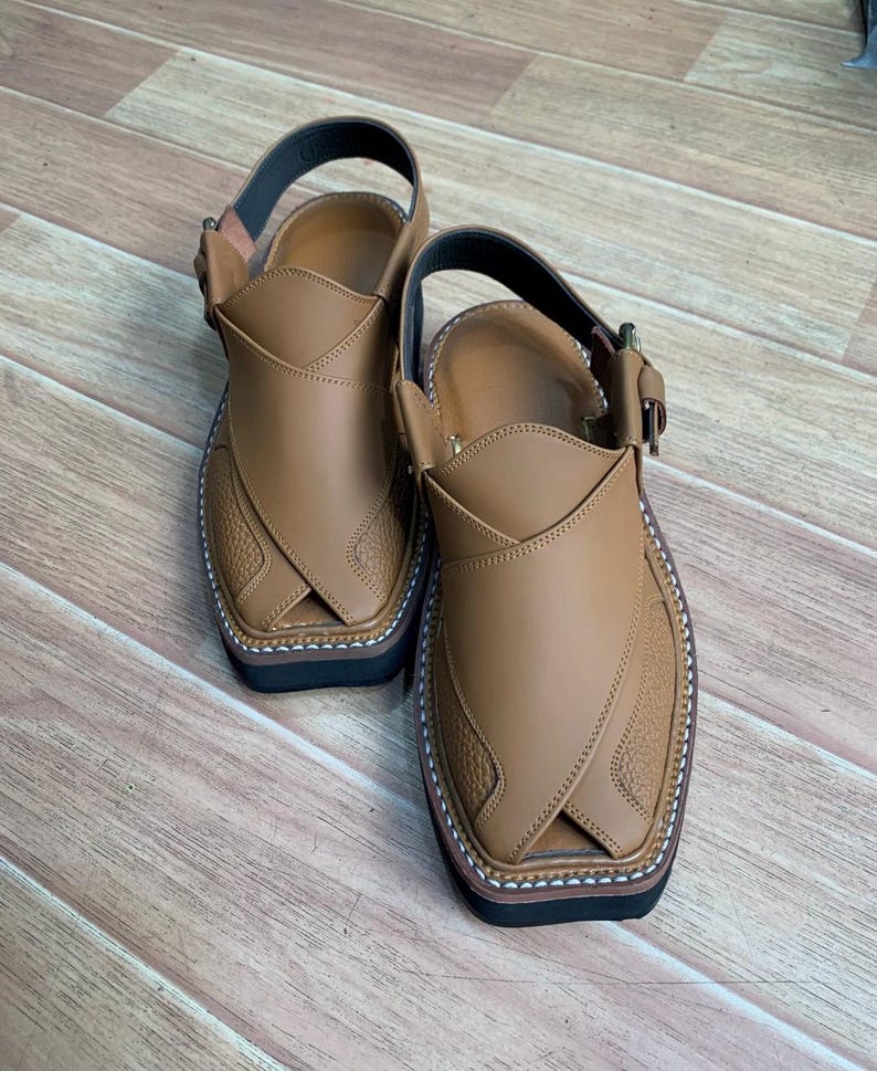 Op de afbeelding: Een paar bruinleren Peshawari-sandalen. De sandalen hebben een bovenwerk met textuur, een enkelband en een zwarte zool. De randen zijn gestikt met witte draad. De sandalen worden gepresenteerd op een houten oppervlak.