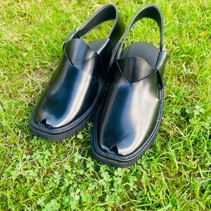 May include: A pair of black leather sandals with a strap across the top and a pointed toe. The sandals are on a green grassy surface.