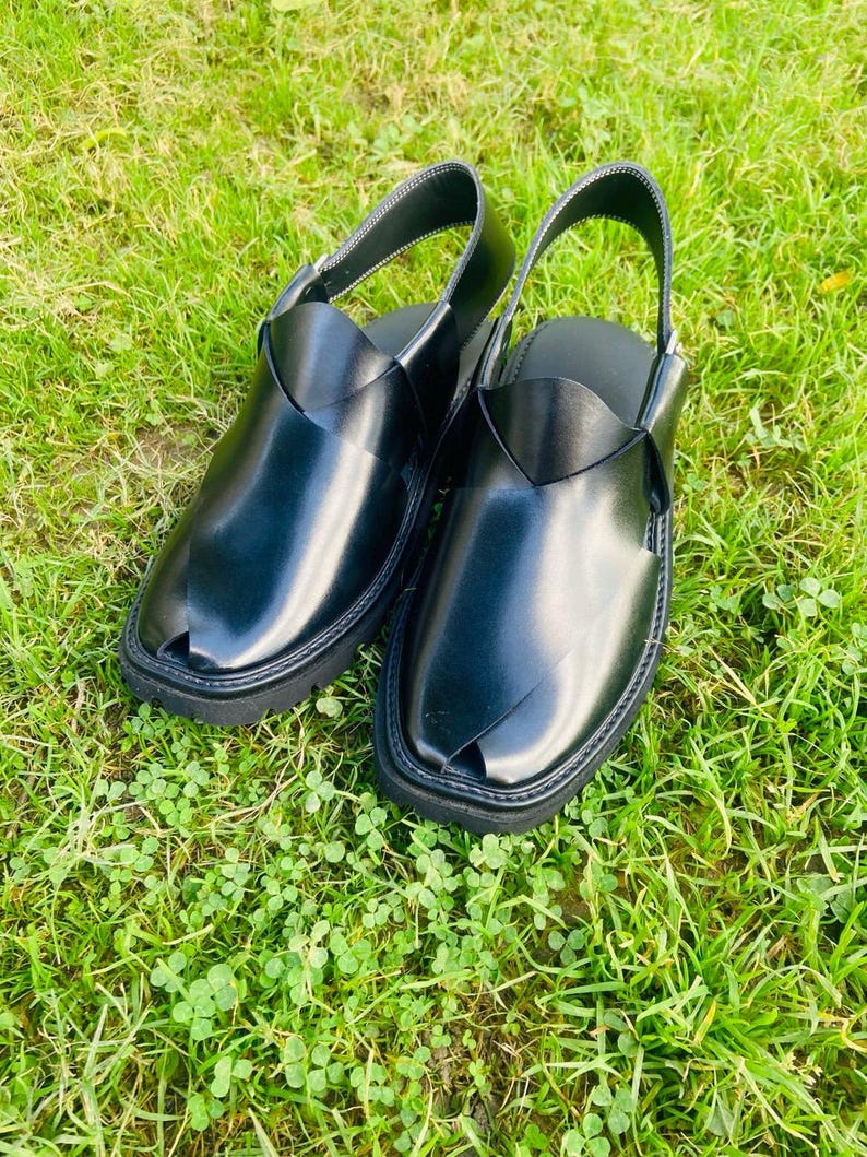 May include: A pair of black leather sandals with a strap across the top and a pointed toe. The sandals are on a green grassy surface.