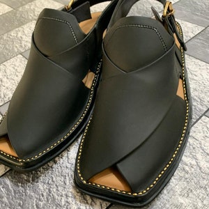 May include: A pair of black leather sandals with a gold buckle and stitching. The sandals have a pointed toe and a strap that goes around the ankle.
