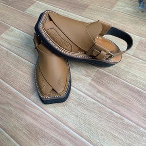 Op de afbeelding: Een paar bruin lederen sandalen met een getextureerd bovenwerk en een zwarte zool. De sandalen hebben een band met een gespsluiting en witte stiksels rond de rand. De sandalen worden gepresenteerd op een houten oppervlak.