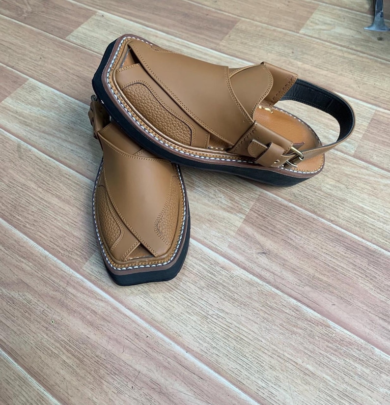 Op de afbeelding: Een paar bruin lederen sandalen met een getextureerd bovenwerk en een zwarte zool. De sandalen hebben een band met een gespsluiting en witte stiksels rond de rand. De sandalen worden gepresenteerd op een houten oppervlak.