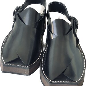 K&ouml;nnte beinhalten: Ein Paar schwarze Ledersandalen mit einem Kreuzriemen-Design. Die Sandalen haben eine braune Sohle und wei&szlig;e N&auml;hte.