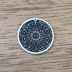 May include: A silver-colored round pendant with a detailed star-like design and Hebrew letters. The pendant has an outer ring with text and is set against a wooden surface.