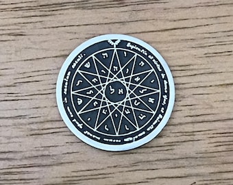 May include: A silver-colored round pendant with a detailed star-like design and Hebrew letters. The pendant has an outer ring with text and is set against a wooden surface.
