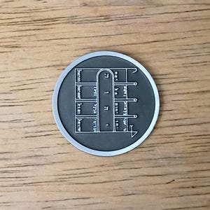 May include: A circular dark gray metal coin featuring a raised silver-toned geometric design. The center displays a vertical arch shape flanked by four horizontal bars on each side, with small etched symbols resembling Hebrew characters within the grid.
