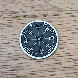 May include: A circular silver-toned metal coin featuring a dark gray recessed center. The surface displays an engraved eight-pointed star design with various symbols at each tip and Hebrew text inscribed along the outer circular border.