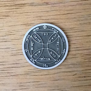 May include: A circular silver-toned metal coin featuring an engraved design of a central cross within a circle, surrounded by a square and additional circular borders. The surface is filled with intricate Hebrew text characters throughout.