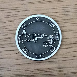 May include: A silver-colored round pendant with Hebrew text and symbols. The design includes a hand pointing, a six-pointed star, and other intricate details. The pendant is set against a wooden surface.