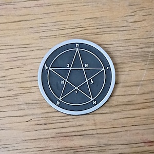 May include: A circular, dark gray metal pendant featuring a pentagram design within a circle. The pentagram is formed by intersecting lines, with Hebrew letters around the star and circle. The pendant is set against a wooden surface.