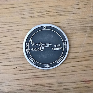 May include: A circular silver-toned metal coin featuring a dark gray recessed center. The design includes a raised hand pointing to the right, Hebrew text, and a small Star of David at the top. The border is smooth and raised, contrasting with the dark background.