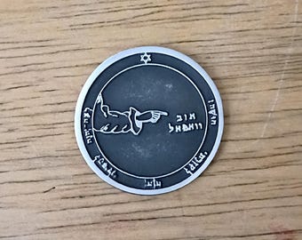 May include: A circular silver-toned metal coin featuring a dark gray recessed center. The design includes a raised hand pointing to the right, Hebrew text, and a small Star of David at the top. The border is smooth and raised, contrasting with the dark background.