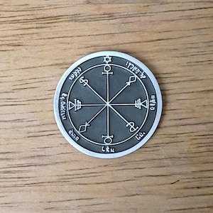 May include: A round, silver-colored pendant with a dark gray center. The pendant features intricate symbols and text around the edge and in the center, with a star at the top. The pendant is on a wooden surface.