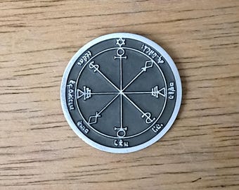 May include: A round, silver-colored pendant with a dark gray center. The pendant features intricate symbols and text around the edge and in the center, with a star at the top. The pendant is on a wooden surface.