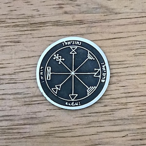 May include: A circular silver-toned metal coin featuring a dark gray recessed center with an engraved eight-pointed star design. The border contains Hebrew text characters etched in a circular pattern around the central geometric symbols.