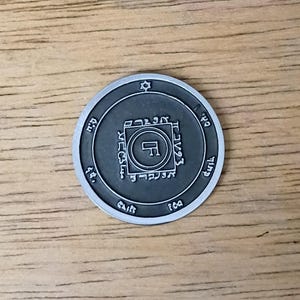 May include: A circular dark grey metal coin featuring a raised silver-toned border and center design. The coin displays a central square enclosing a symbol, surrounded by Hebrew-style characters, a small star at the top, and additional symbols.
