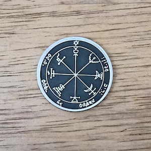 May include: A round, silver-colored pendant with a dark gray center. The center features a star-like design with symbols and text around the edge. The pendant is set against a wooden surface.