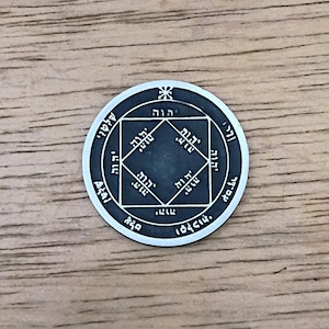 May include: A circular silver-toned metal coin featuring an engraved square design at the center. The surface displays intricate Hebrew characters arranged within the square and along the outer circular border, with a small star symbol at the top.