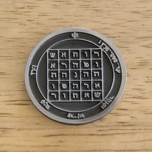 May include: A circular, silver-colored metal pendant with an engraved square grid of Hebrew letters. The pendant has an outer ring with additional Hebrew text and a small cross-like symbol at the top. The pendant is displayed on a wooden surface.