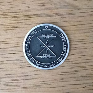 May include: A silver-colored circular pendant with Hebrew text and geometric designs. The pendant features a Star of David at the top and intricate symbols within a central circle. The background is a light brown wooden surface.
