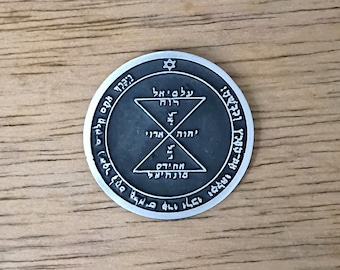 May include: A silver-colored circular pendant with Hebrew text and geometric designs. The pendant features a Star of David at the top and intricate symbols within a central circle. The background is a light brown wooden surface.