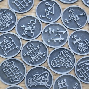 K&ouml;nnte beinhalten: Ein Satz von 16 runden, silberfarbenen Spielsteinen mit wei&szlig;en, komplizierten Designs. Jeder Spielstein zeigt ein anderes Symbol oder Muster.
