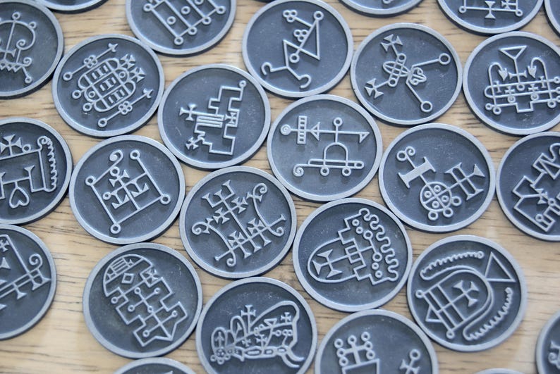 K&ouml;nnte beinhalten: Ein Satz von 16 runden, silberfarbenen Spielsteinen mit wei&szlig;en, komplizierten Designs. Jeder Spielstein zeigt ein anderes Symbol oder Muster.