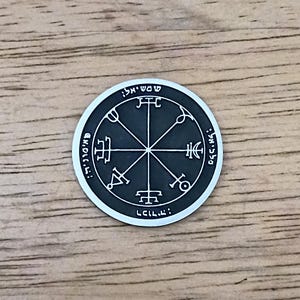 May include: A silver-colored round pendant with a black center featuring intricate symbols and text. The design includes a central star-like pattern with lines radiating outwards, surrounded by Hebrew text. The pendant is set against a wooden surface.