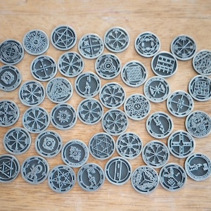 May include: A collection of silver-colored, circular tokens with intricate, engraved designs. The tokens feature various geometric patterns, symbols, and illustrations, arranged on a wooden surface. The designs include stars, lines, and other esoteric imagery.