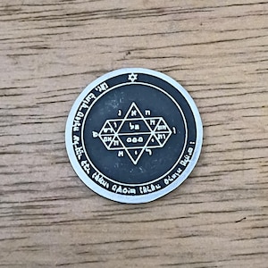 May include: A circular silver-toned metal medallion featuring a central six-pointed star design filled with Hebrew characters. The outer border of the coin-shaped object is inscribed with additional Hebrew text and a small star symbol at the top.