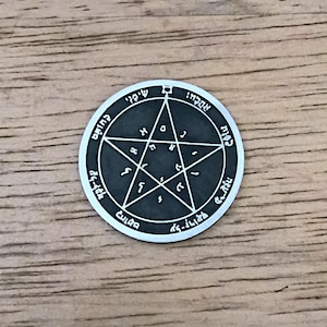 May include: A silver-colored round pendant with a dark gray center. The pendant features a pentagram design with Hebrew text around the edge and within the star. The pendant is set against a wooden background.