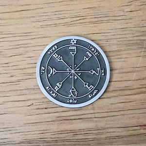 May include: A silver-colored circular pendant with intricate engravings and Hebrew text. The design features a central star, arrows, and symbols within a circular border. The pendant is displayed on a wooden surface.