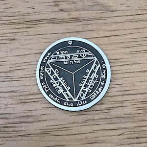 May include: A silver-colored round pendant with Hebrew text and geometric designs. The pendant features a central triangle and surrounding text, with a star at the top. The pendant is displayed on a wooden surface.
