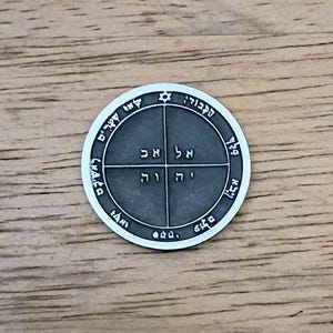 May include: A circular silver-toned metal medallion featuring a dark gray center divided into four quadrants by a cross. Each quadrant contains Hebrew characters. The outer border displays additional Hebrew text and a small six-pointed star symbol.