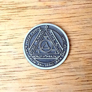 May include: A circular silver-toned metal coin featuring an engraved design of a central triangle within a larger triangle, surrounded by a circular border filled with intricate symbols and characters, with a small star at the top apex.