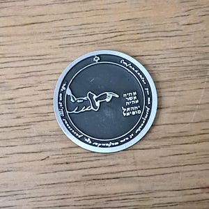 May include: A silver-colored round pendant with a dark gray center. The center features an illustration of a hand reaching towards a dove, with Hebrew text. The outer rim has additional text. The pendant is on a wooden surface.