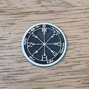 May include: A silver-colored, circular pendant with intricate engraved symbols and text. The design features a central star, radiating lines, and various shapes within a circular border. The pendant is set against a wooden surface.