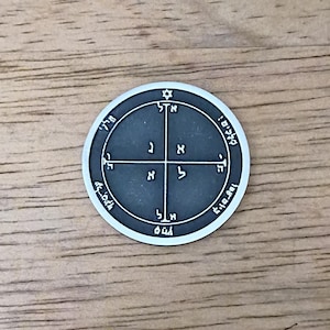 May include: A circular silver-toned metal medallion featuring a dark gray recessed center. The surface displays a cross design with Hebrew characters in each quadrant and along the outer border, topped with a small six-pointed star symbol.