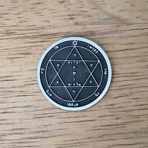 May include: A circular dark gray metal medallion featuring a raised silver-toned hexagram at its center. The design includes Hebrew characters inscribed within the star and along the outer circular border, topped with a small six-pointed star symbol.