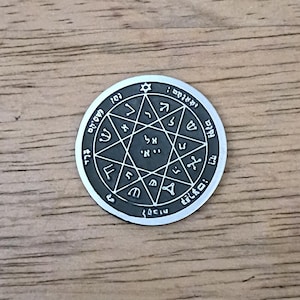 May include: A circular silver-toned metal coin featuring an engraved star pattern with twelve points. The center contains Hebrew characters, surrounded by additional Hebrew script and symbols arranged in a circular border around the perimeter.