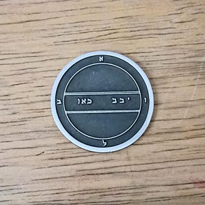 May include: A silver-colored circular pendant with a dark gray center. The pendant features Hebrew text and symbols within concentric circles. The background is a light brown wooden surface.