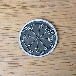 May include: A circular silver-toned metal coin featuring a dark gray recessed center. The surface displays a central eight-pointed star design with intricate symbols at each tip and various etched characters arranged around the outer border.