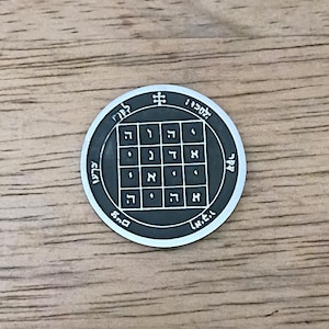 May include: A circular dark grey metal coin featuring a central five-by-five grid containing Hebrew characters. The outer border of the coin displays additional Hebrew text and a small cross-like symbol at the top, all in a raised silver finish.