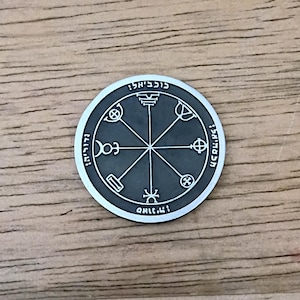 May include: A circular silver-toned metal coin featuring a dark gray recessed center with an engraved eight-pointed star design. The outer rim displays embossed Hebrew characters and various symbolic markings arranged in a radial pattern.