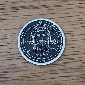 May include: A silver-colored round coin with an engraved design. The coin features a bearded face, Hebrew text, and surrounding script. A six-pointed star is at the top. The coin is set against a wooden surface.