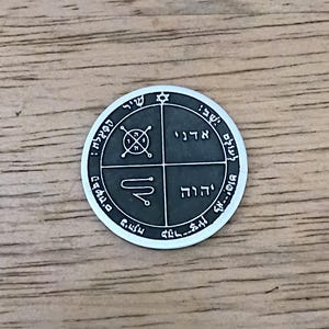 May include: A silver-colored, circular pendant with engraved symbols and Hebrew text. The pendant features a Star of David at the top and is divided into quadrants with various symbols and inscriptions. The background is a wooden surface.