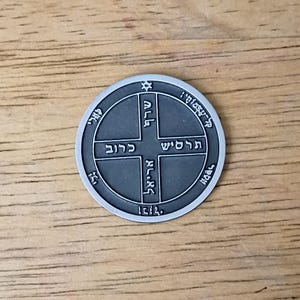 May include: A circular silver-toned metal medallion featuring a raised cross design. The surface is engraved with Hebrew characters within the cross and along the outer circular border. A small star symbol is positioned at the top of the inner circle.