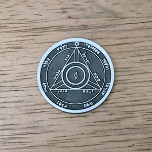 May include: A circular silver-toned metal coin featuring an engraved design of a central flame inside a triangle, surrounded by a circle with Hebrew text and a small star at the top. The coin has a dark, recessed background with raised silver details.