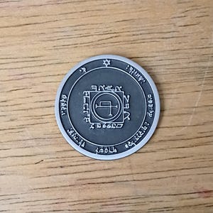 May include: A circular silver-toned metal coin featuring a dark gray recessed center. The design includes a central square frame enclosing a sigil, surrounded by a ring of engraved symbols and text, with a small six-pointed star at the top edge.