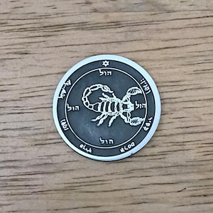 May include: A circular silver-toned metal coin featuring a raised scorpion graphic in the center. The coin has a dark grey recessed background and a border inscribed with Hebrew characters and a small Star of David symbol at the top position.